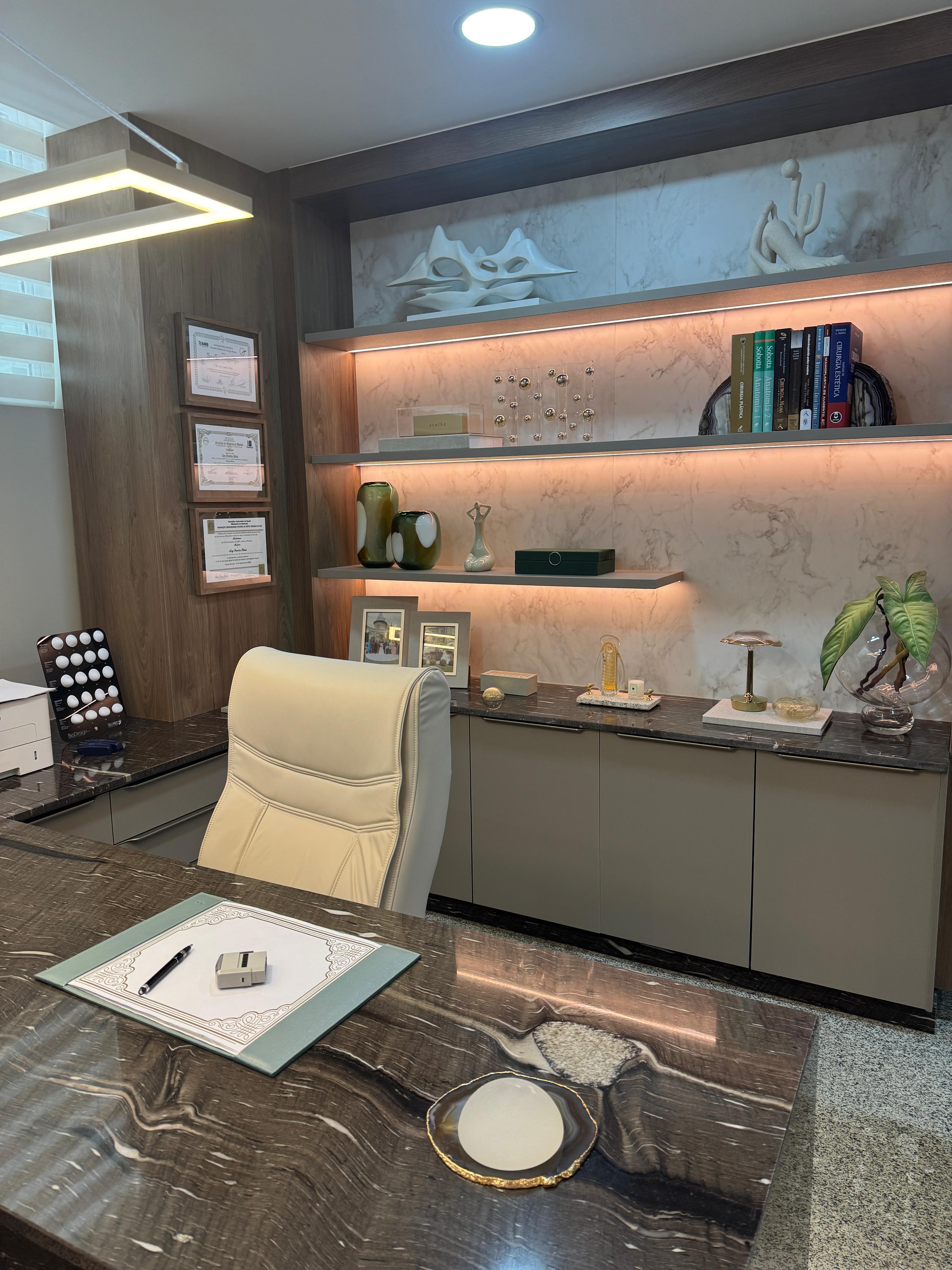 Sala de consulta, com mesa de mármore escuro, poltrona clara e estantes iluminadas com livros e objetos