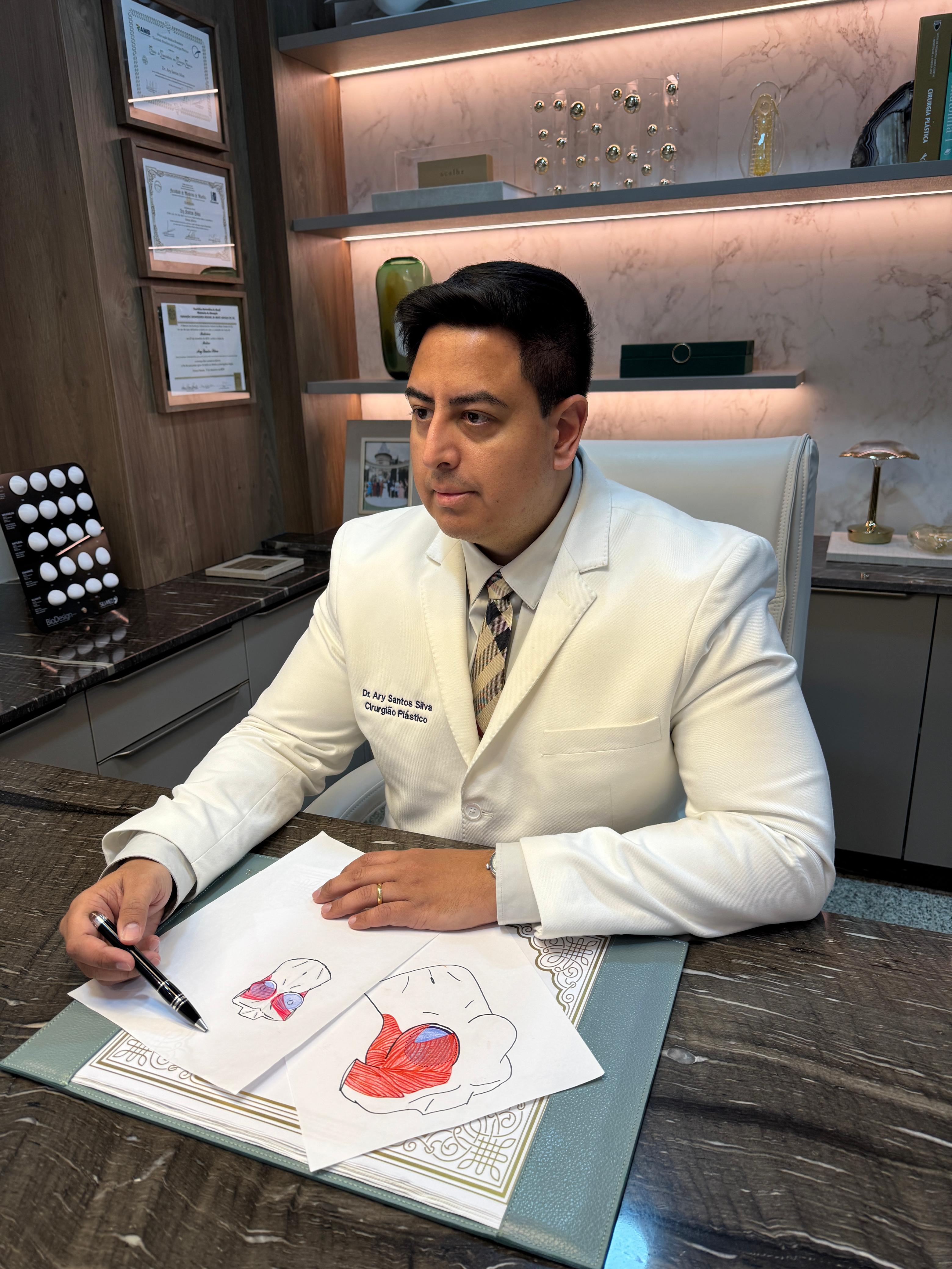 Dr. Ary Santos Silva, de jaleco, sentado à mesa do consultório, com desenhos de planejamento cirúrgico sobre a mesa